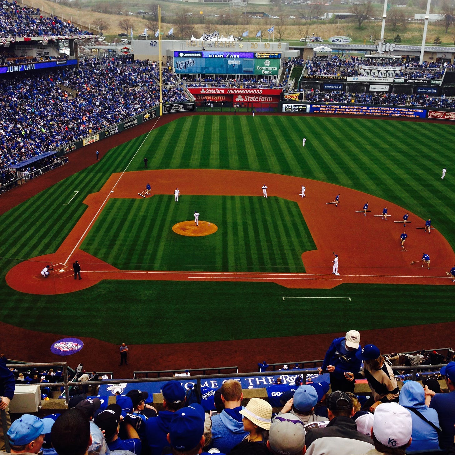 A baseball diamond, grass surrounding the dirt infield, from the point of view of the crowd high up along the first base side.