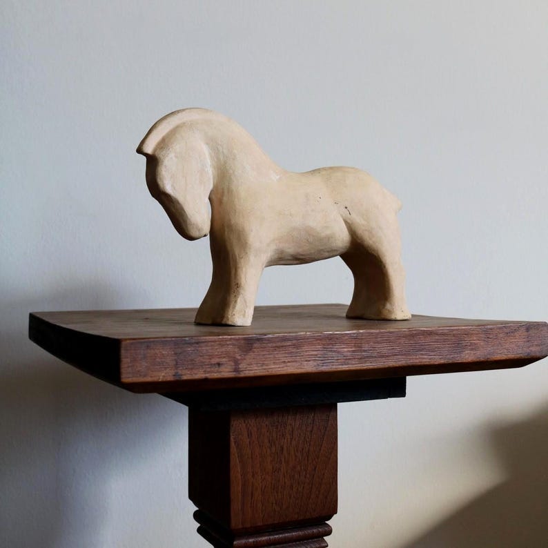 May include: A light beige, carved wooden horse sculpture. The horse has a simple, stylized form with a curved neck and a slightly lowered head. It stands on a dark brown wooden shelf, which is supported by a dark brown pedestal. May include: A light beige, carved wooden horse sculpture. The horse has a simple, stylized form with a curved neck and a slightly lowered head. It stands on a dark brown wooden shelf, which is supported by a dark brown pedestal.