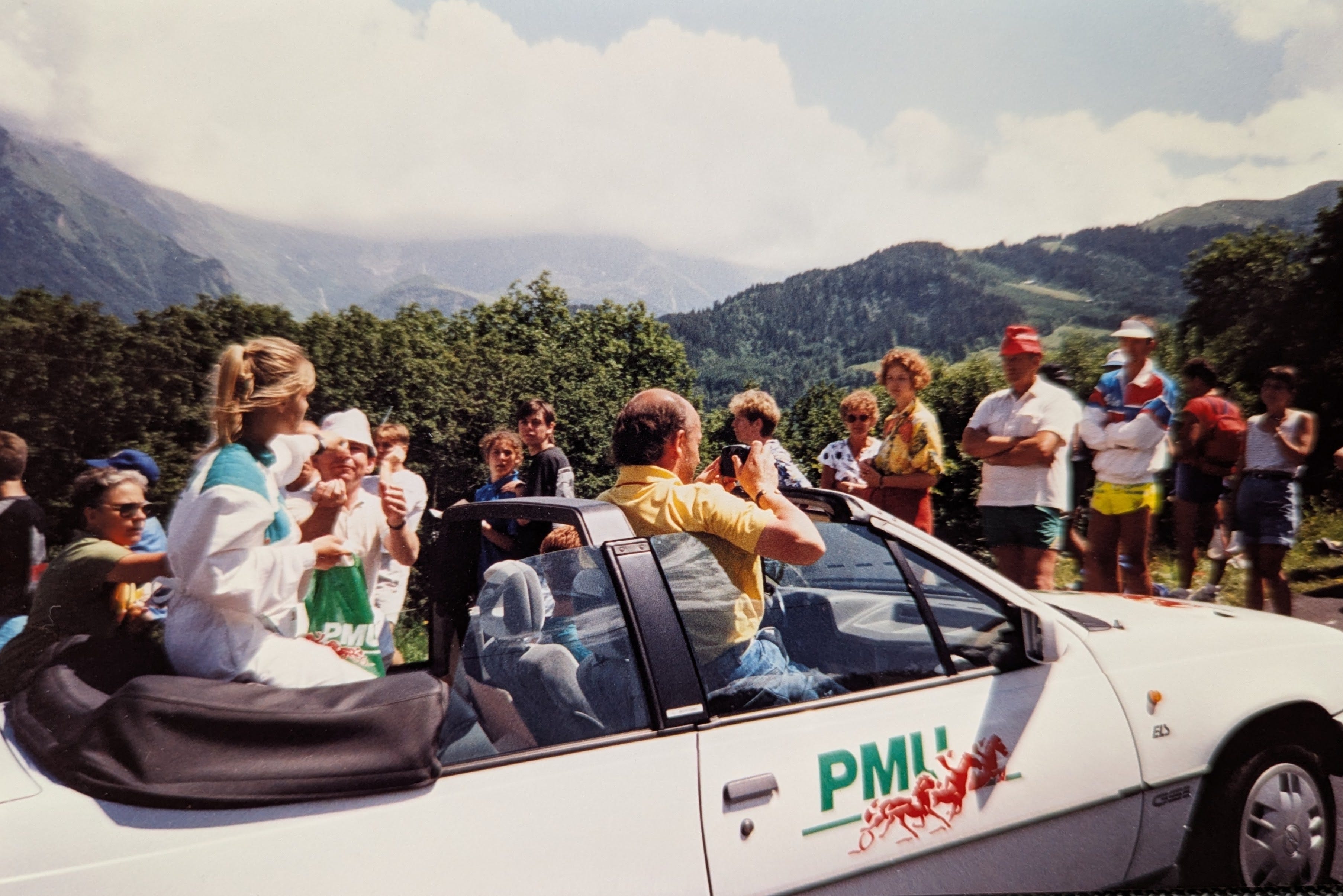 A slightly elevated outdoor shot captures a white convertible car driving uphill with a crowd of people gathered around it under a partly cloudy sky. The car is angled from the front passenger side, with its top down.  In the front passenger seat, a person with short brown hair, wearing a yellow collared shirt, is lifting himself above the windshield and taking a picture with a camera held to their eye, facing towards the right side of the frame. In the back seat on the left, a blonde-haired woman in a white and teal top is seated on the folded convertible top, her head above the rollbar  The car has a green "PMI" logo with two red stylized horse figures on its front passenger door.  Numerous spectators are visible around the car. On the left side of the frame, several people are standing near the car. On the right side of the car, a larger group of people of varying ages and attire are standing and looking towards the general direction of the car or further into the background.  The background features lush green trees and a mountainous landscape under a bright sky with scattered clouds. The mountains are hazy in the distance. The overall impression is of a public event, possibly a race or parade, where spectators are eagerly watching.