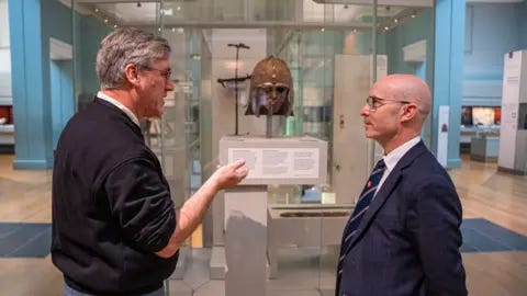 Kent County Council Two men talking to one another while stand in front of a decorative metal helmet in a museum case