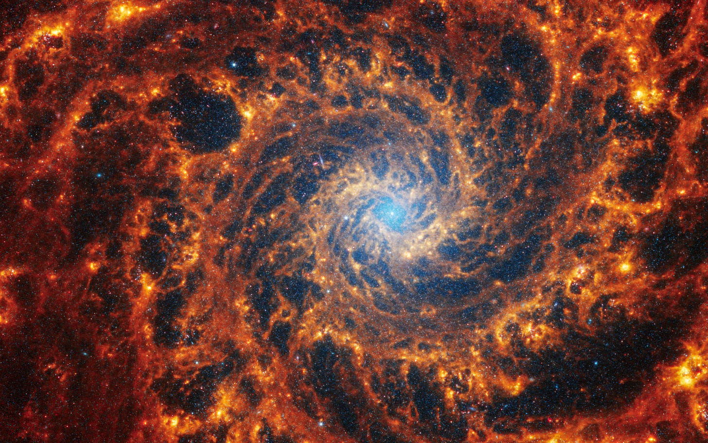Webb’s image of NGC 628 shows a densely populated face-on spiral galaxy anchored by its central region, which has a light blue haze that takes up about a quarter of the view. In this circular core is the brightest blue area. Within the core are populations of older stars, represented by many pinpoints of blue light. Spiny spiral arms made of stars, gas, and dust also start at the center, largely starting in the wider area of the blue haze. The spiral arms extend to the edges, rotating counterclockwise. The spiraling filamentary structure looks somewhat like a cross section of a nautilus shell. The arms of the galaxy are largely orange, ranging from dark to bright orange. Scattered across the packed scene are some additional bright blue pinpoints of light, which are stars spread throughout the galaxy. In areas where there is less orange, it is darker, and some dark regions look more circular. A prominent dark “bubble” appears to the top left of the blue core. And a wider, elliptical “bubble” to the bottom right. Webb’s image of NGC 628 shows a densely populated face-on spiral galaxy anchored by its central region, which has a light blue haze that takes up about a quarter of the view. In this circular core is the brightest blue area. Within the core are populations of older stars, represented by many pinpoints of blue light. Spiny spiral arms made of stars, gas, and dust also start at the center, largely starting in the wider area of the blue haze. The spiral arms extend to the edges, rotating counterclockwise. The spiraling filamentary structure looks somewhat like a cross section of a nautilus shell. The arms of the galaxy are largely orange, ranging from dark to bright orange. Scattered across the packed scene are some additional bright blue pinpoints of light, which are stars spread throughout the galaxy. In areas where there is less orange, it is darker, and some dark regions look more circular. A prominent dark “bubble” appears to the top left of the blue core. And a wider, elliptical “bubble” to the bottom right.
