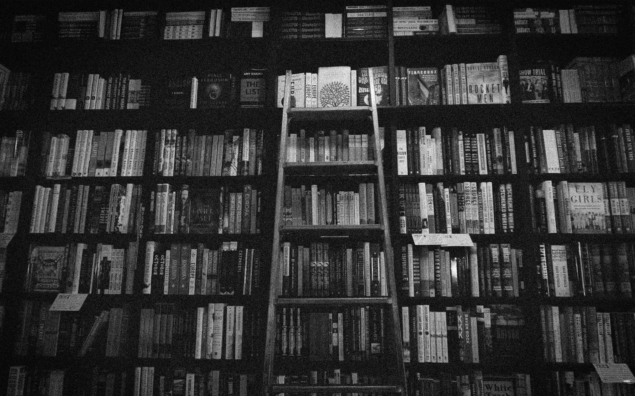 Photograph depicts hundreds of books on floor to ceiling bookshelves, with a wooden ladder placed against it. Photograph depicts hundreds of books on floor to ceiling bookshelves, with a wooden ladder placed against it.