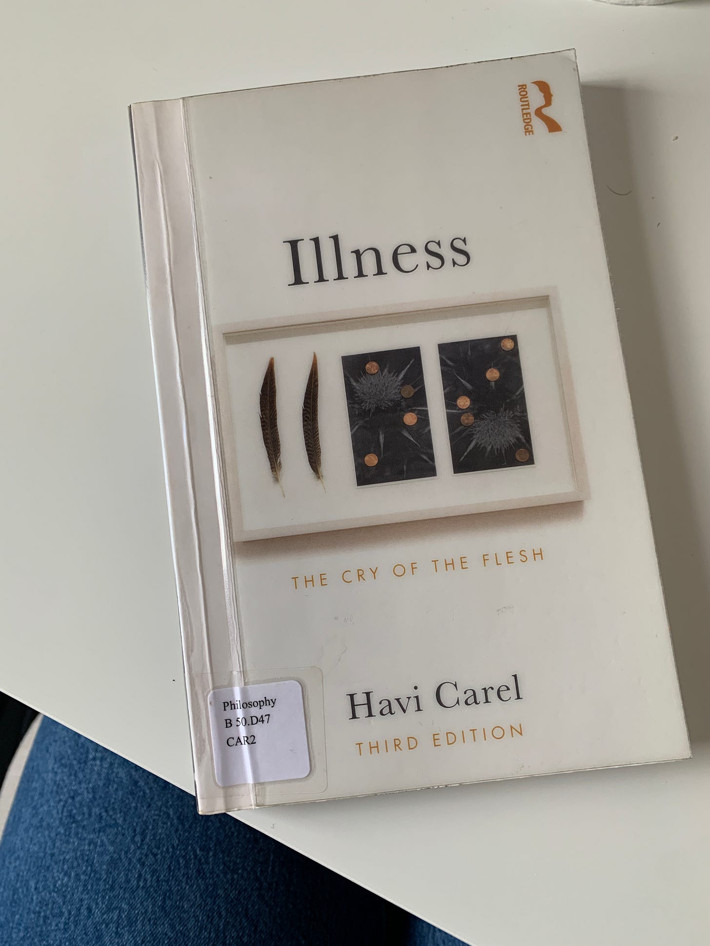 A copy of Havi Carel's book Illness laid closed on a white desk. The book cover is white with blacktext and the image of a framed artwork that consists of two long feathers and two abstract images that are black rectangles with gold circles and grey scratch markings like a burst of fireworks.