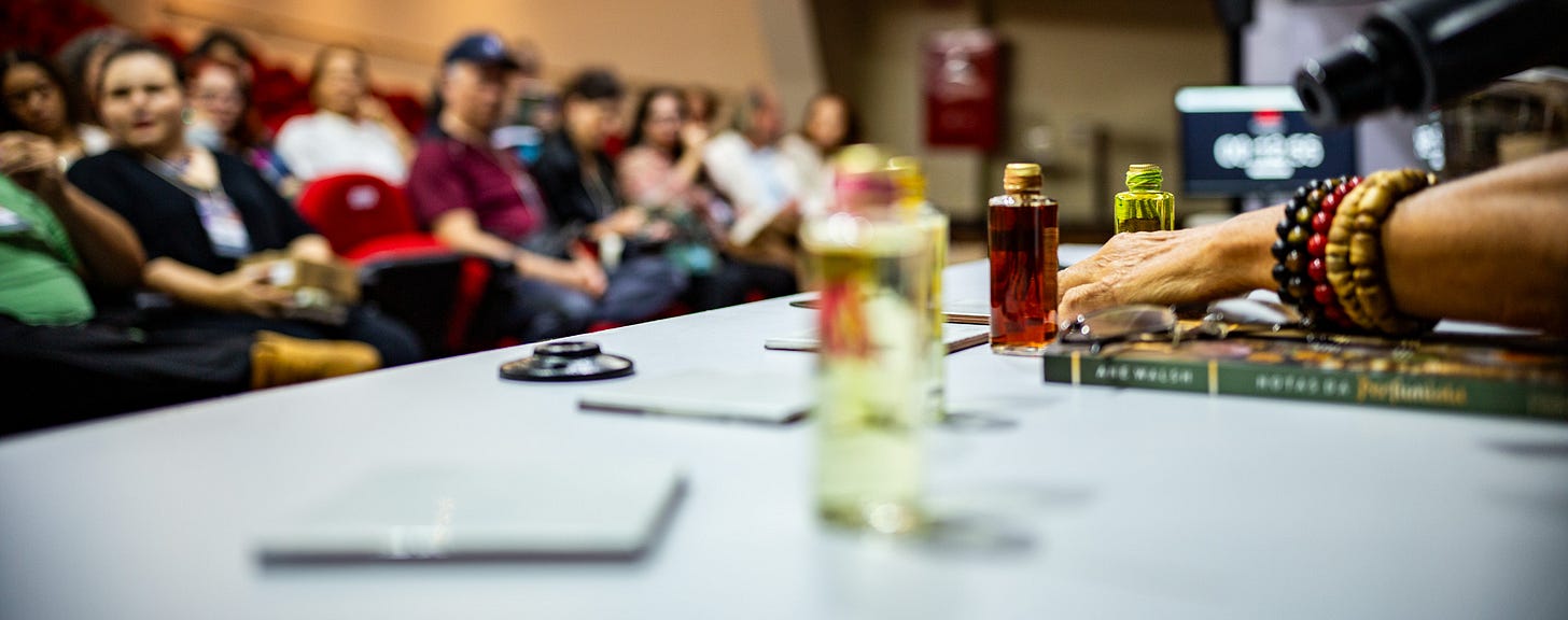 Ane Walsh’s hand with beaded bracelets gesturing over bottles of natural perfume oils on a presentation table with a blurred audience in the background