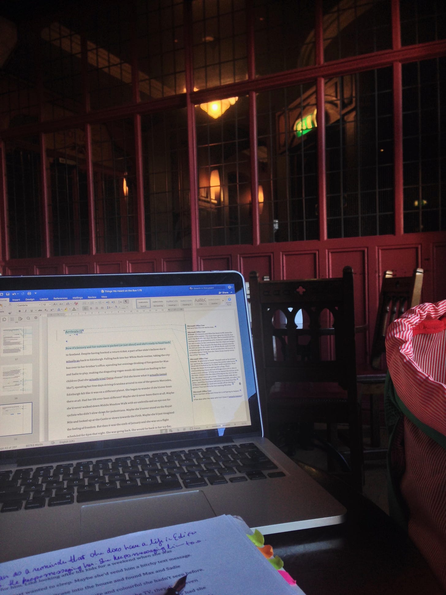 A Victorian space with a red wall of windows and wood chairs in the background. In the foreground is an open laptop on a table, a printed manuscript with handwritten notes, and a fountain pen and backpack.