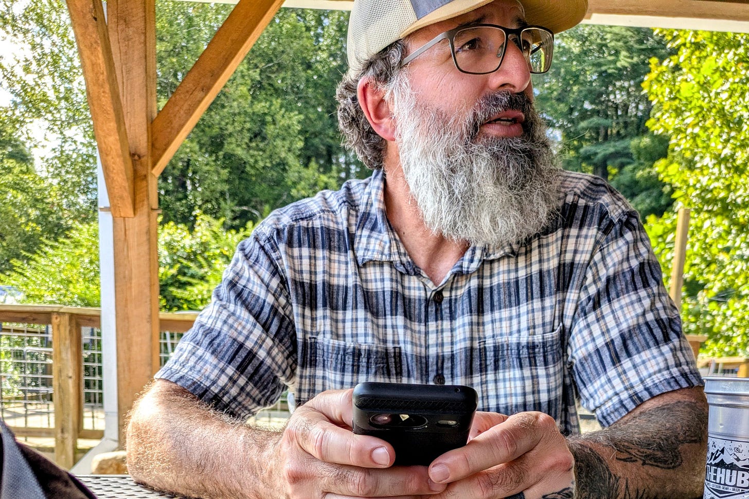 The author sits outdoors under a wooden shelter, holding a smartphone with both hands while looking off into the distance. He wears glasses, a baseball cap, and a plaid shirt, with trees and bright summer greenery in the background.