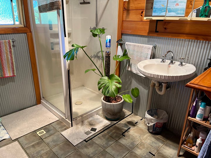 four photos, the first one shows a dated bathroom with dark floor tiles and a shower with a big monstera plant in front of it. The next photo shows the same space with new whitish grey floor tiles just laid. The third photo is an outdoor shot with a pile of garbage that includes: an old toilet, ripped up tiles and other debris from a housing demolition. The final photo is of a front porch with a yellow screen door in the center. On one side is a tile cutting machine on a drop cloth with many other tools of the trade and on the opposite side is a pile of wood trim and boxes of tiles.