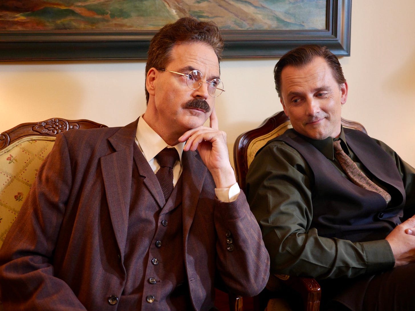two men sit next to each other under a painting, one in full three-piece suit and spectacles, the other in shirt and waistcoat
