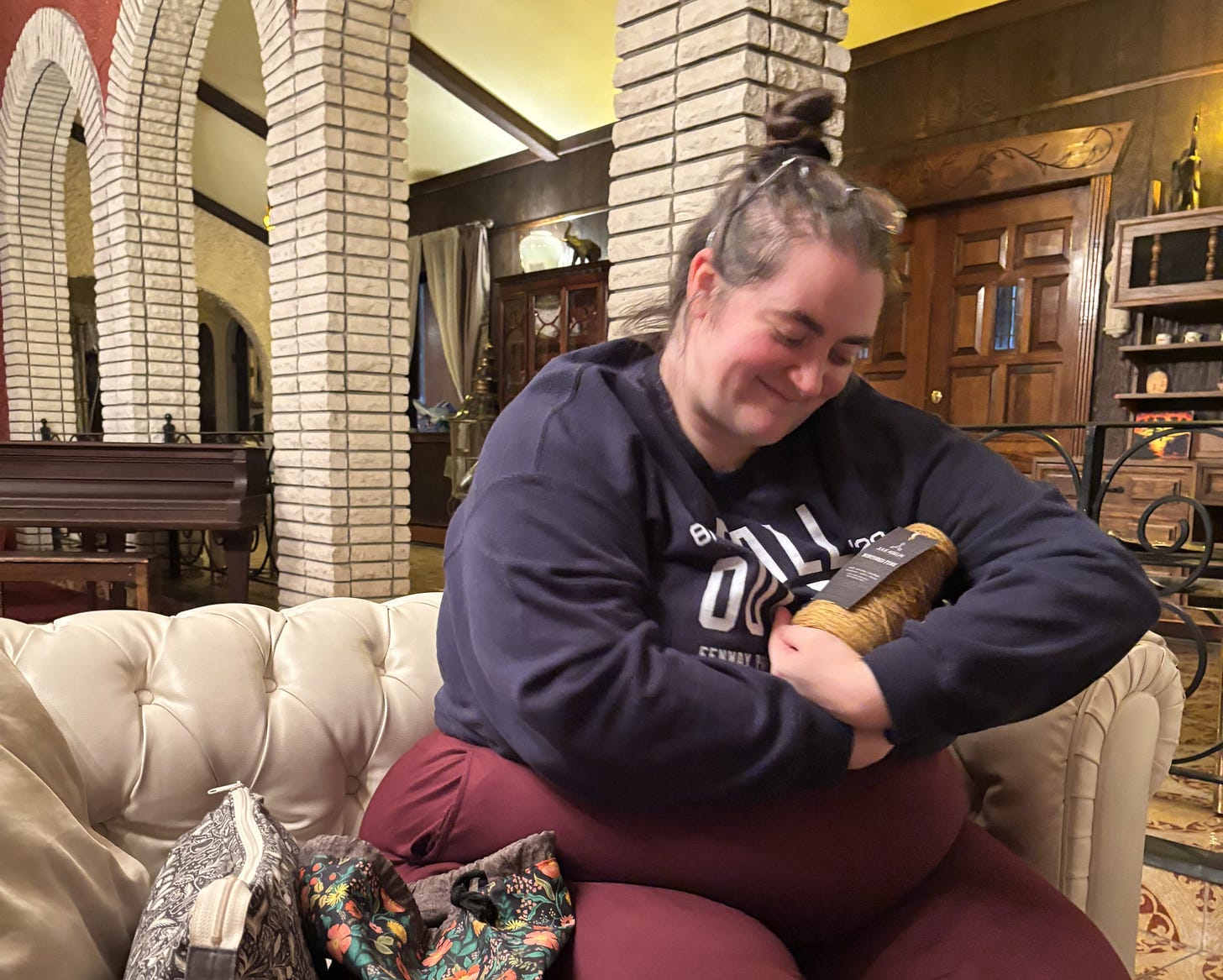 A fat white woman sits on a white couch with two project bags beside her. She is cradling a cone of golden yellow yarn. She has a smile on her face.