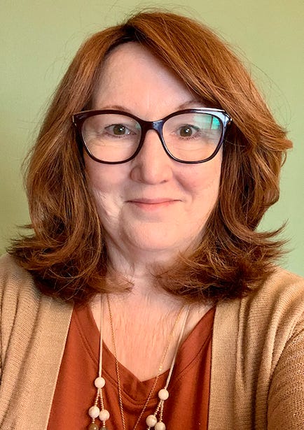 Photograph of the author, who has red hair, dark glasses, and is wearing a rusty-colored shirt with tan cardigan.