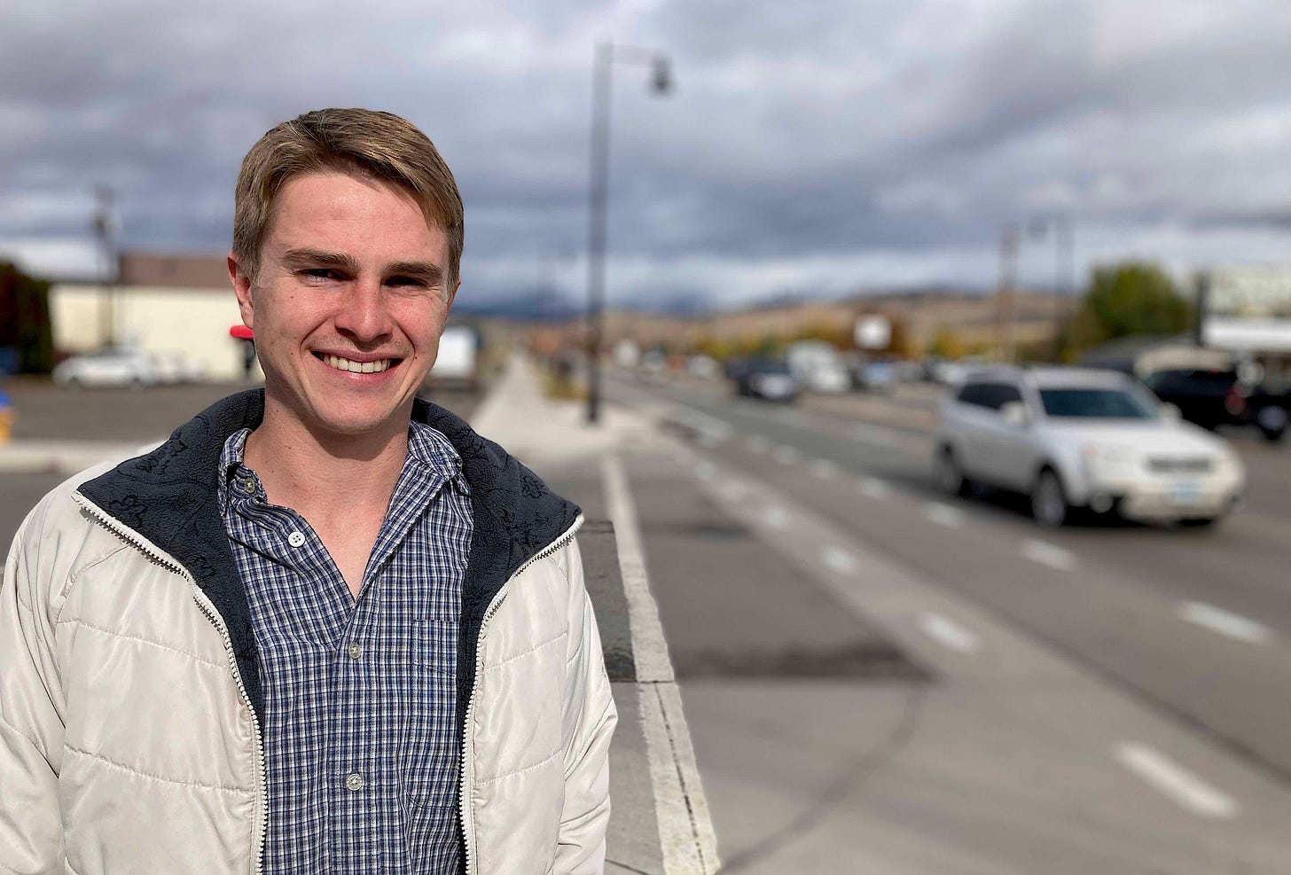 man in coat and checkered shirt smiling on street corner