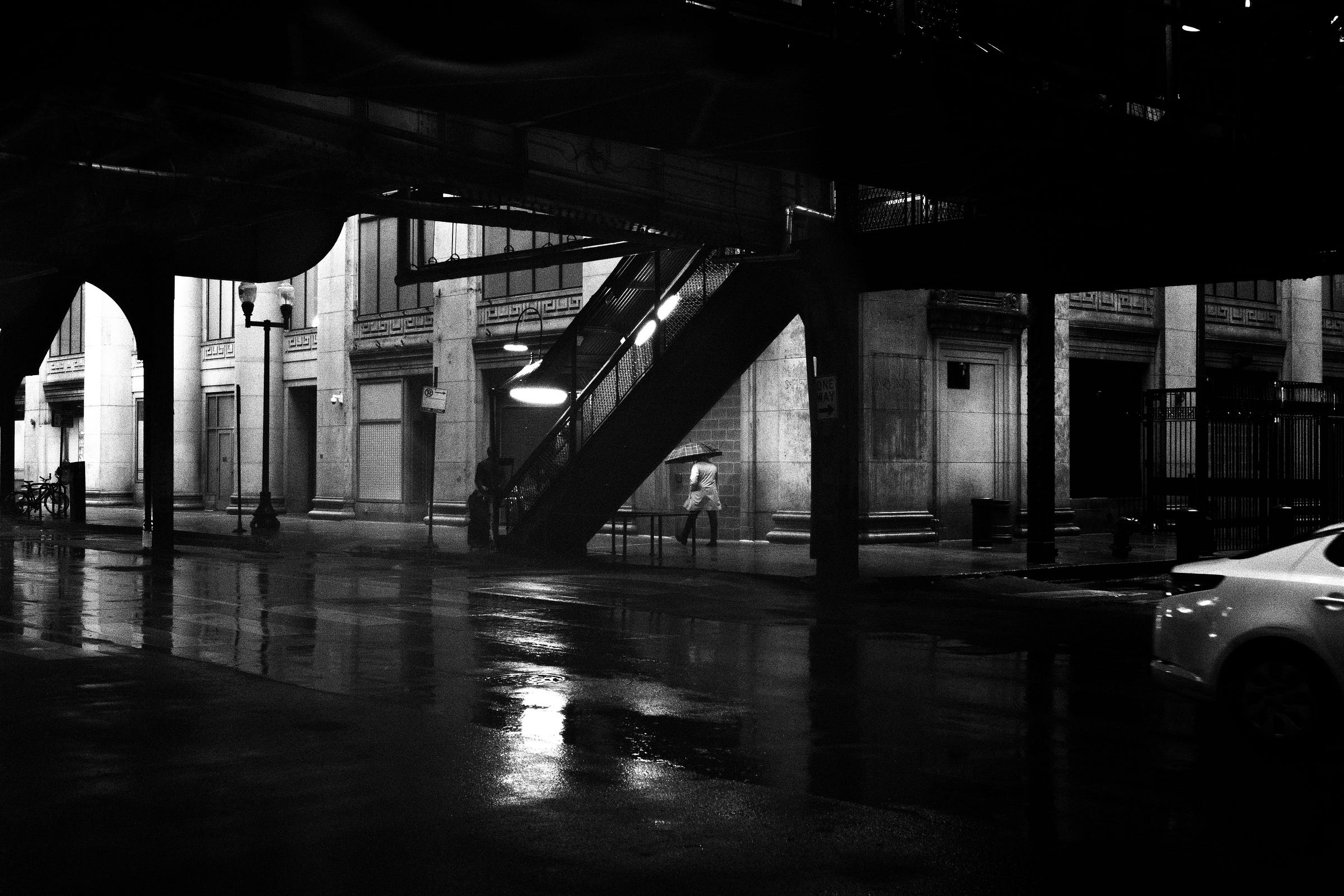 A solitary figure with an umbrella walks beneath an elevated train platform on a rainy night, their reflection shimmering on the wet pavement as streetlights cast a moody glow. A solitary figure with an umbrella walks beneath an elevated train platform on a rainy night, their reflection shimmering on the wet pavement as streetlights cast a moody glow.