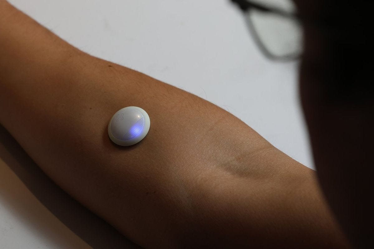 A Biolinq Shine sensor worn on the forearm, a small white disc with a glowing blue light