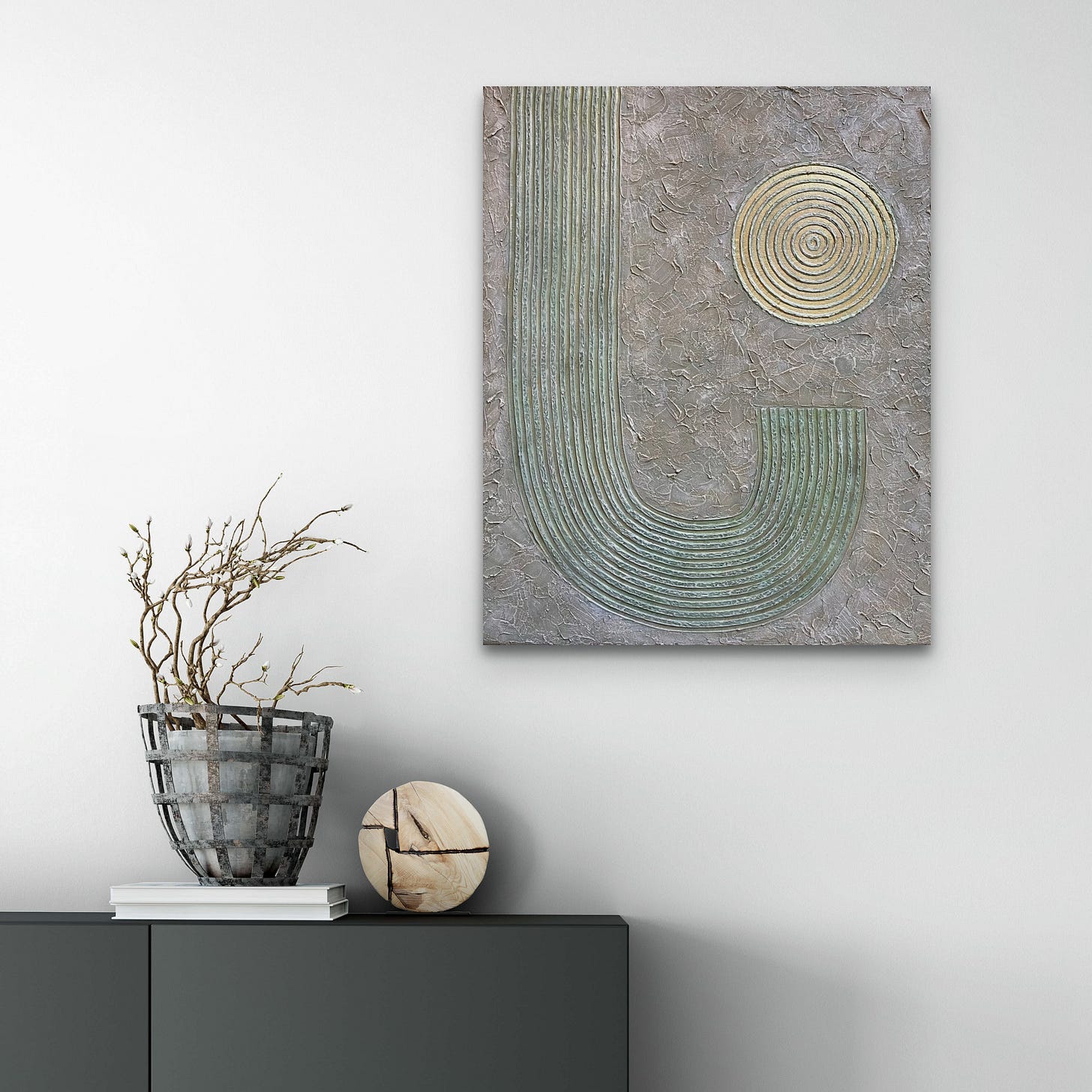 Textured abstract artwork in muted earth tones featuring a raised U-shape pattern and a concentric circular motif on a rough, stone-like surface. The painting hangs on a minimalist wall above a dark cabinet with a sculptural twig arrangement in a rustic metal basket and a round, cracked wooden object.