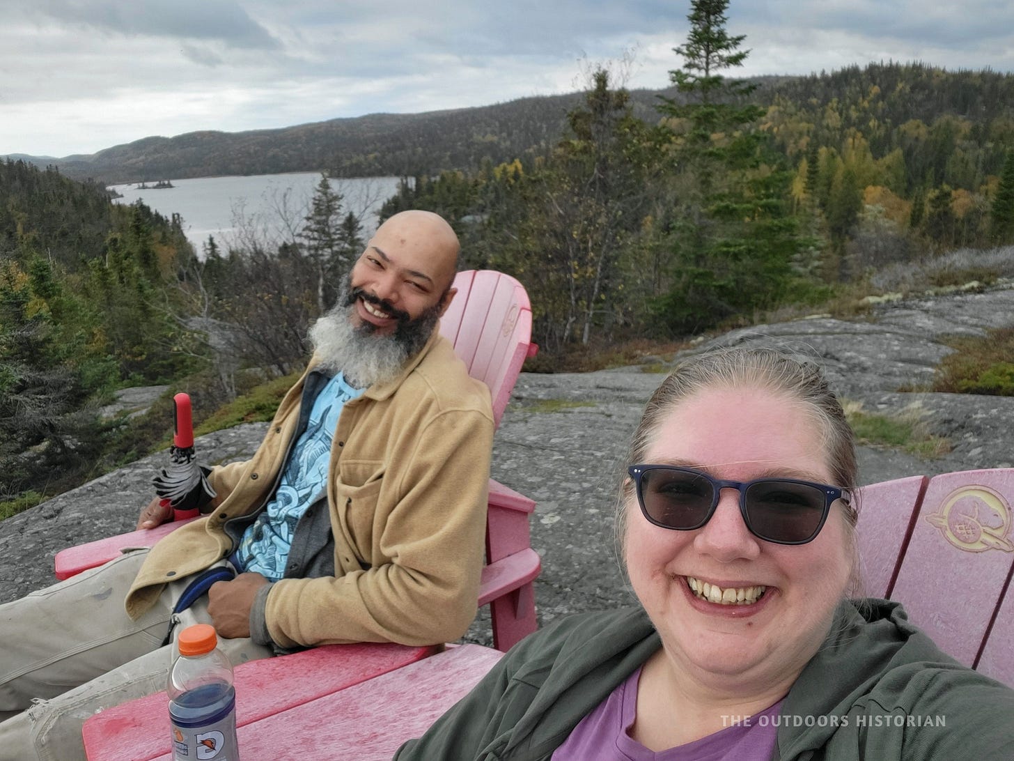 A selfie of Emily and Eugene sitting side-by-side in the red Parks Canada chairs. They are smiling, bundled in warm hiking clothes, with the beautiful, rocky expanse of Hattie Cove behind them.