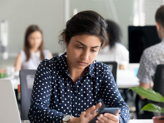 Serious pensive worker sitting at desk typing message using smartphone.