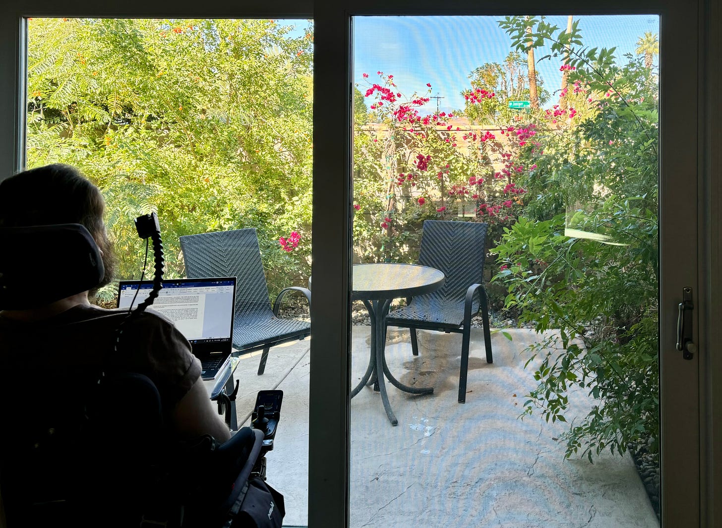 Eric sits in his motorized wheelchair and gazes out of a wide sliding glass door. Resting atop his wheelchair tray is a PC laptop open to an unfinished Word document. The patio beyond has a small glass-top table and two chairs. Orange trumpet vines and magenta flowers grow up all around the patio.