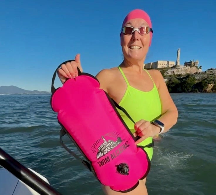 A smiling swimmer wearing a neon green swimsuit and pink swim cap holds a bright pink Swim Alcatraz swim buoy while in open water near Alcatraz Island. The buoy is designed for visibility and flotation, making it an essential safety tool for open water swimming. A smiling swimmer wearing a neon green swimsuit and pink swim cap holds a bright pink Swim Alcatraz swim buoy while in open water near Alcatraz Island. The buoy is designed for visibility and flotation, making it an essential safety tool for open water swimming.
