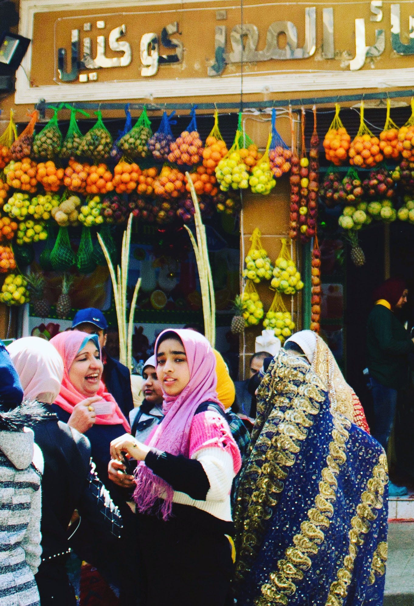 A group of women and girls in bright headscarves laugh and talk outside a juice bar on Al-Muizz Street in Cairo, beneath hanging nets of oranges, apples, and stalks of sugarcane.