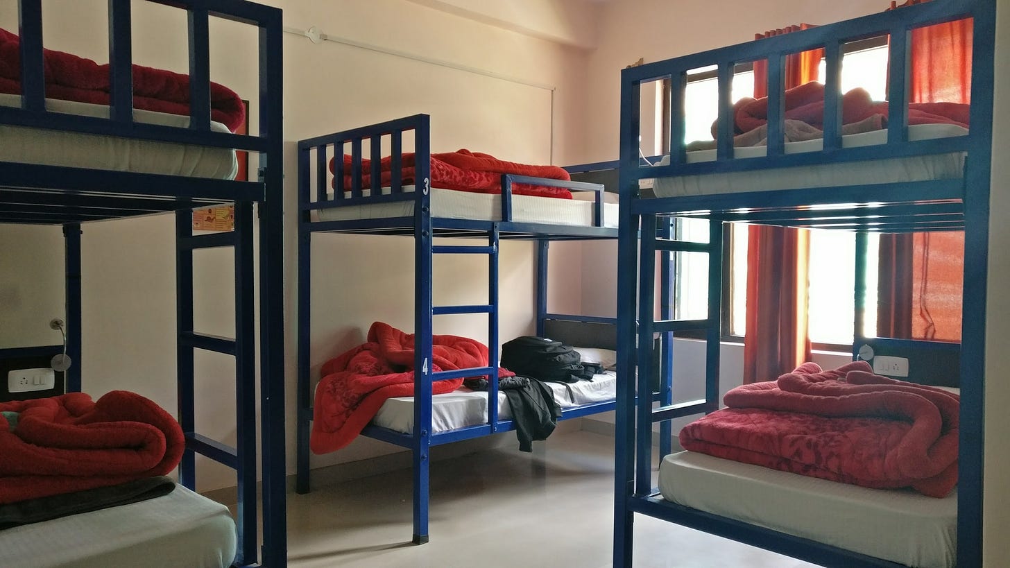 A hostel dorm room with blue metal bunk beds, red blankets folded on each bed, a backpack on one lower bunk.
