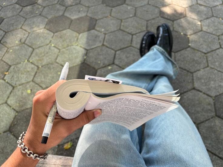 This may contain: a person sitting on a bench holding a book with a pen in their left hand This may contain: a person sitting on a bench holding a book with a pen in their left hand