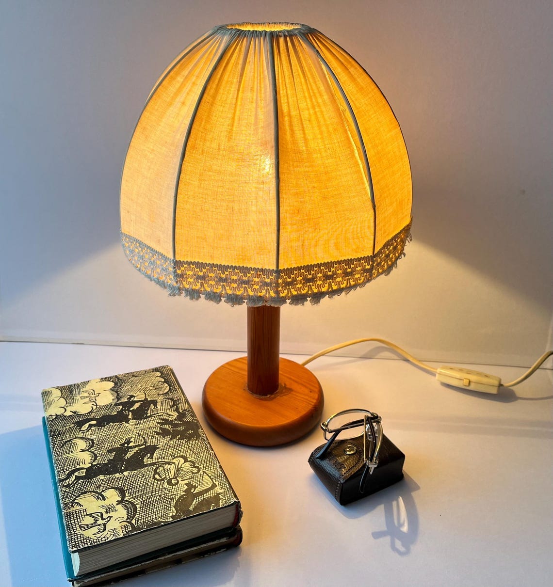 May include: A vintage table lamp with a yellow lampshade and a wooden base. The lampshade has a scalloped edge with lace trim. A book with a black and white cover and a pair of glasses in a small case are next to the lamp. May include: A vintage table lamp with a yellow lampshade and a wooden base. The lampshade has a scalloped edge with lace trim. A book with a black and white cover and a pair of glasses in a small case are next to the lamp.