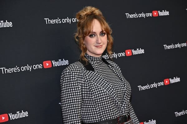 Brittany Broski, wearing a black-and-white checked outfit, stands for a portrait in front of a screen with the YouTube logo. 