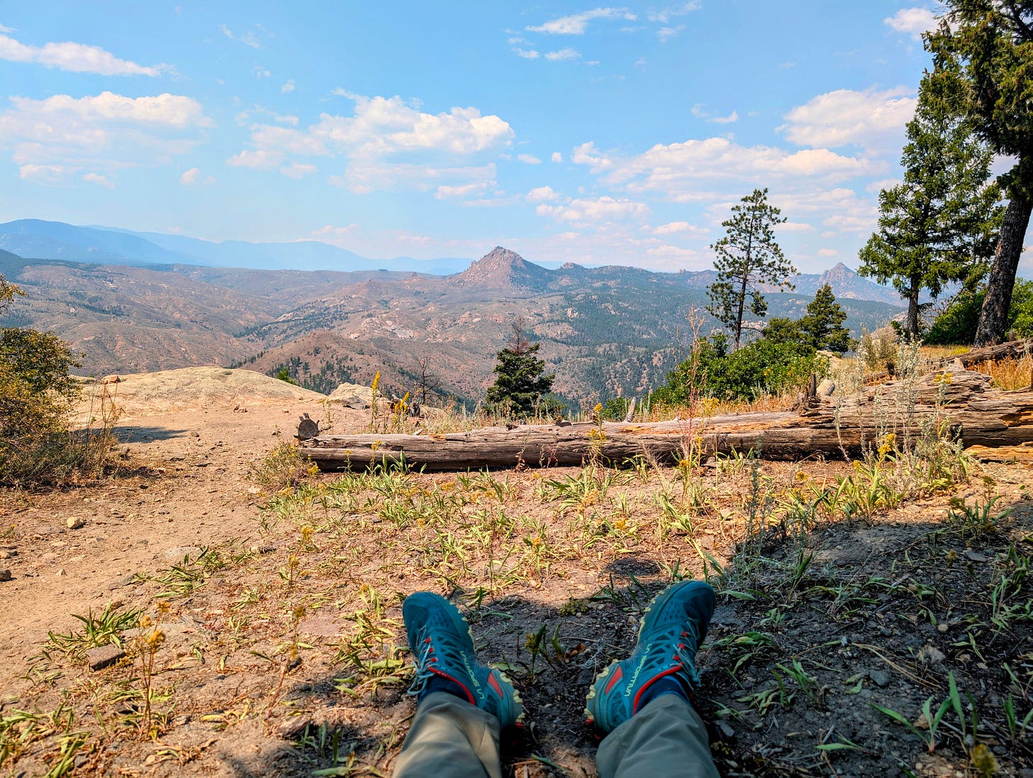 Hiker’s legs and blue trail runners stretched out during a break, overlooking a wide mountain valley under a blue sky. Hiker’s legs and blue trail runners stretched out during a break, overlooking a wide mountain valley under a blue sky.