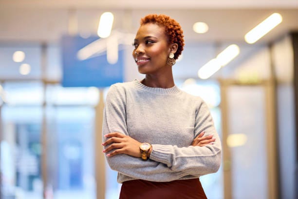 Confident Professional Woman Looking Aside Stylish African American woman with crossed arms looking away in a blurred office setting. She wears a gray sweater, maroon skirt, and gold watch. confidence stock pictures, royalty-free photos & images