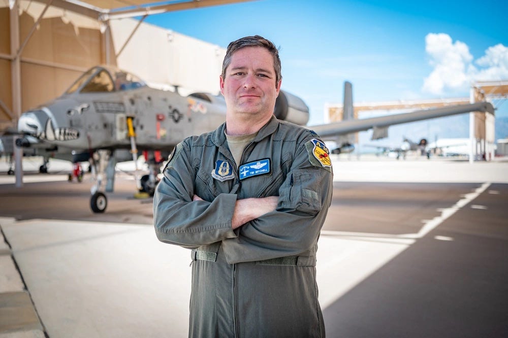 A middle aged man in a flight suit stands with arms crossed. An A-10 is in the background. 