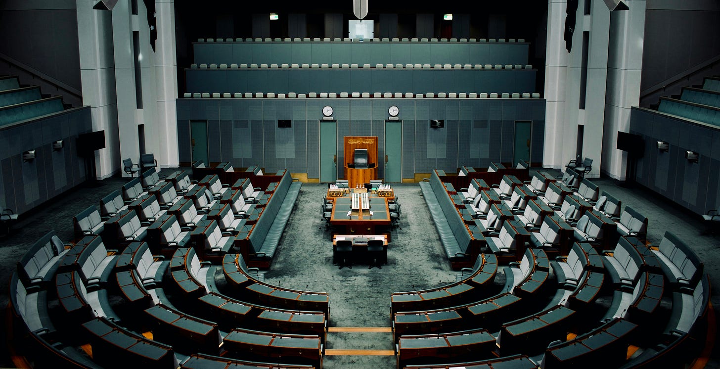 A wide-angle photograph of an empty parliamentary debating chamber, with curved rows of unoccupied seats facing a central podium, lit by cool overhead lighting.
