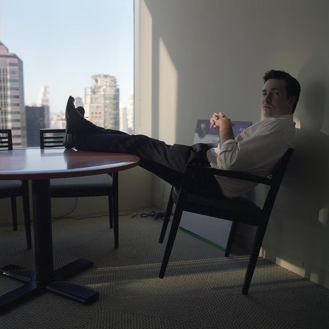 white guy in business casual leaning back in a chair in front of a window; outside there are skyscrapers white guy in business casual leaning back in a chair in front of a window; outside there are skyscrapers