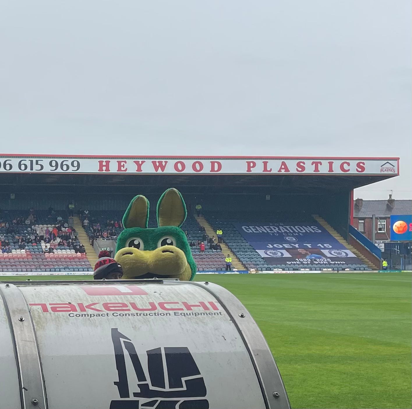 Rochdale's mascot (who looks like a green rabbit, but is, apparently, a dragon) peeps over the top of the away dugout at Spotland football ground.