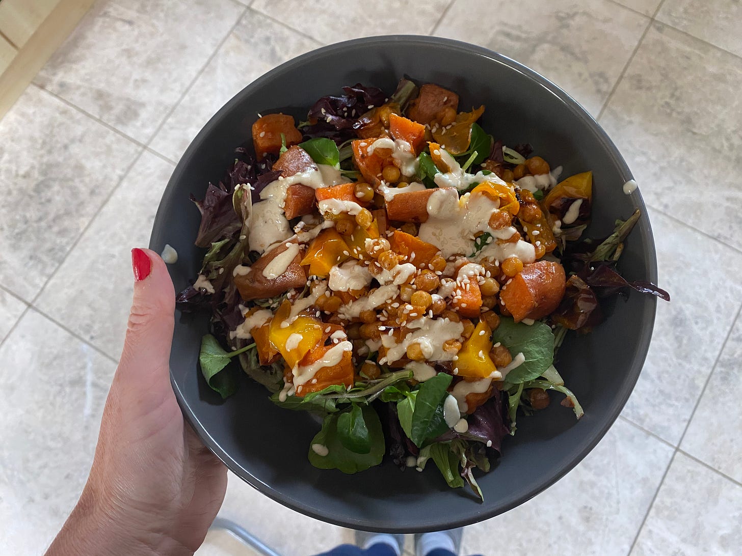Left hand holding a grey bowl. In the bowl is a salad with roasted squash, leaves, beans and a creamy sauce