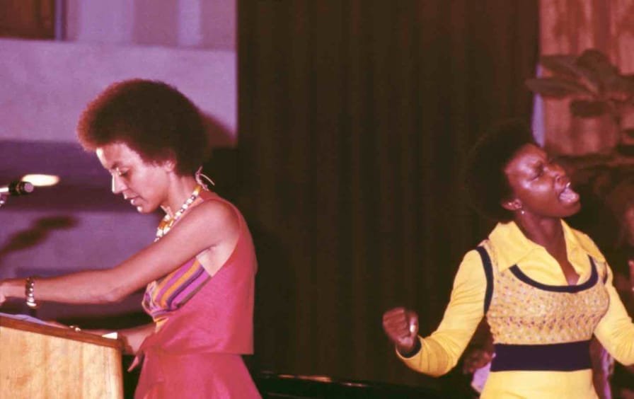 Nikki Giovanni standing by the lectern and woman singing during a performance at The Phillis Wheatley Poetry Festival., 1973.