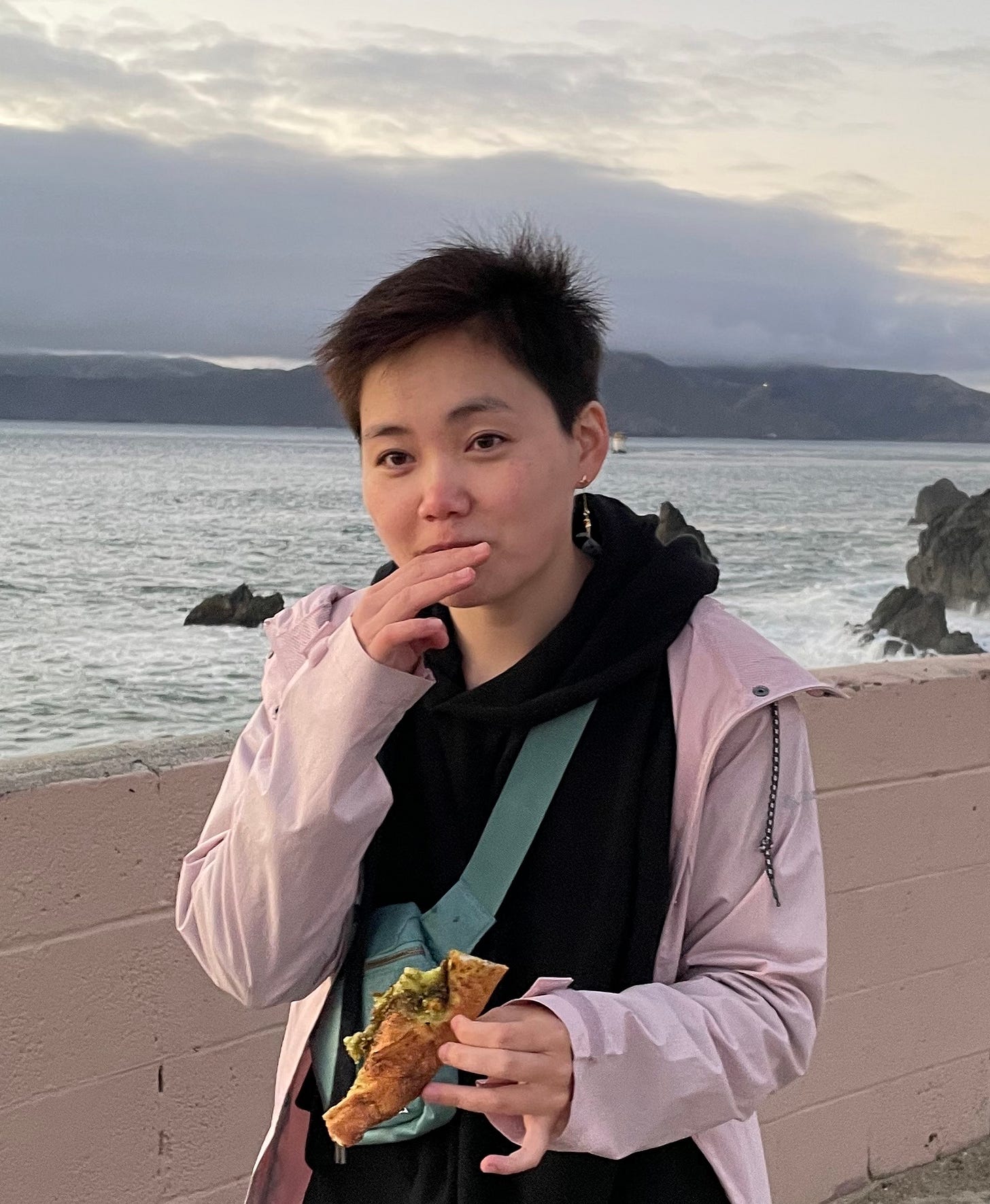 a person with short black hair standing outside and eating a slice of pizza. behind them is a seashore and cloudy sky. a person with short black hair standing outside and eating a slice of pizza. behind them is a seashore and cloudy sky.