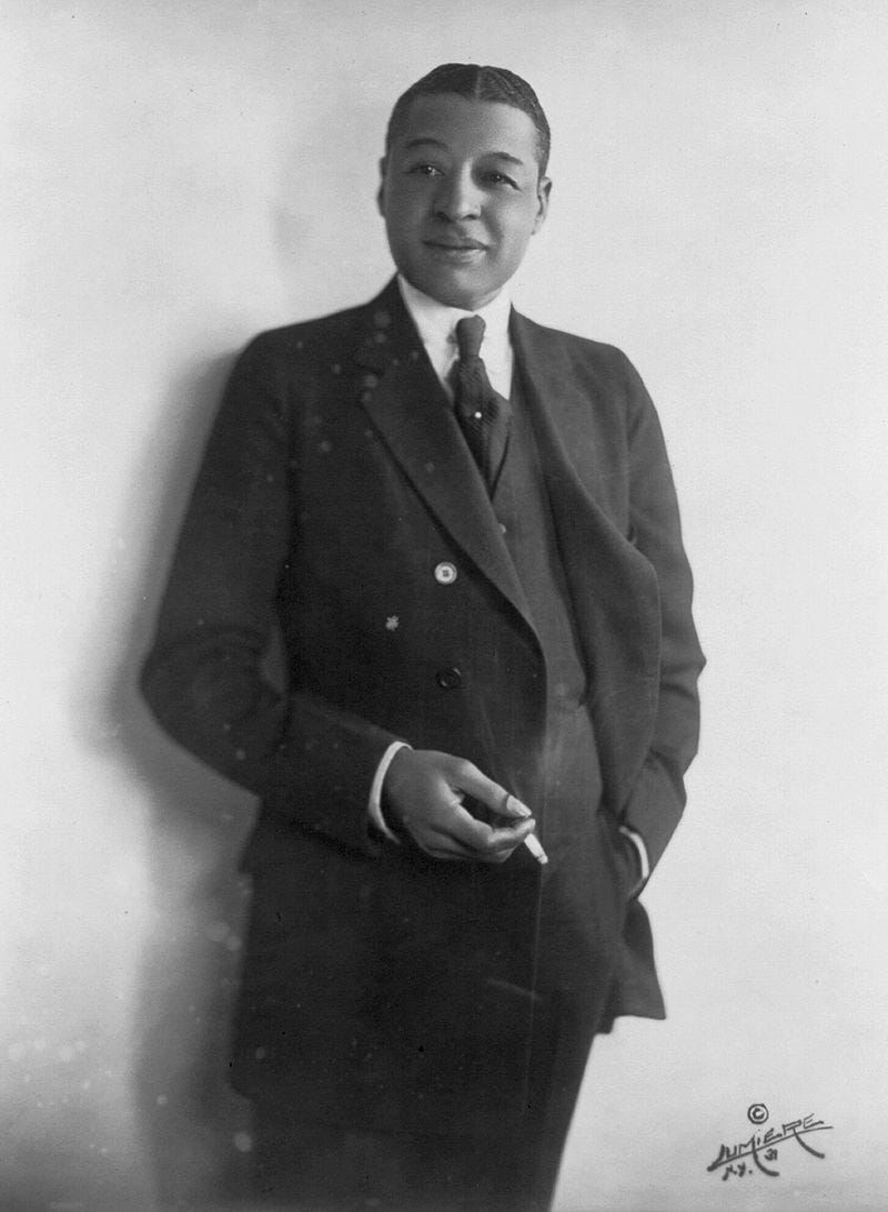 black and white Photo portrait of Vaudeville star Bert Williams, seen standing, with a lit cigarette in right hand.