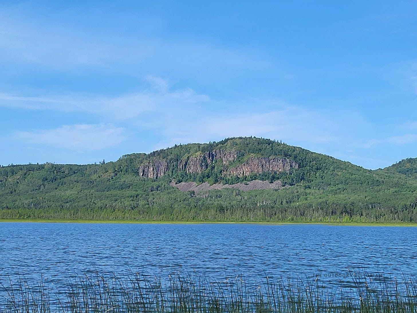 Steep, vertical rock cliffs rising sharply from the talus slopes above a forested bluff that slopes to the wild rice beds along the shore at the west end of Whitefish Lake.
