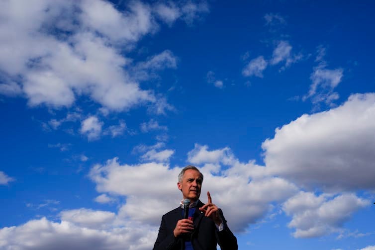 A man with short grey hair speaks into a microphone with a large blue sky as a backdrop. A man with short grey hair speaks into a microphone with a large blue sky as a backdrop.