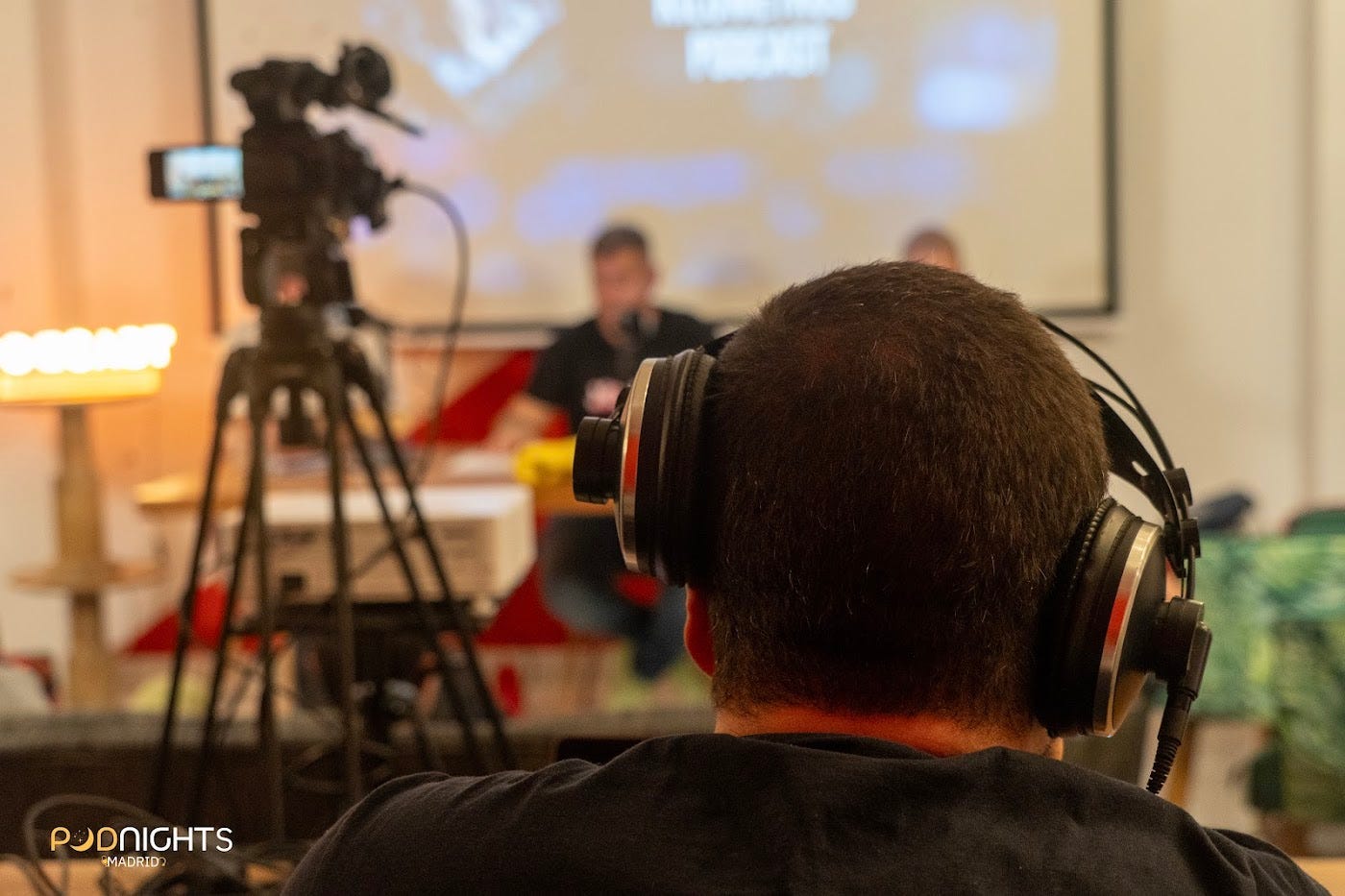 Persona con auriculares observando y escuchando la grabación de un podcast en directo. En primer plano se ve la parte trasera de su cabeza, mientras al fondo aparecen una cámara de vídeo sobre trípode y dos personas participando en una mesa de grabación frente a una pantalla. La imagen está tomada durante un evento de Podnights Madrid. Persona con auriculares observando y escuchando la grabación de un podcast en directo. En primer plano se ve la parte trasera de su cabeza, mientras al fondo aparecen una cámara de vídeo sobre trípode y dos personas participando en una mesa de grabación frente a una pantalla. La imagen está tomada durante un evento de Podnights Madrid.