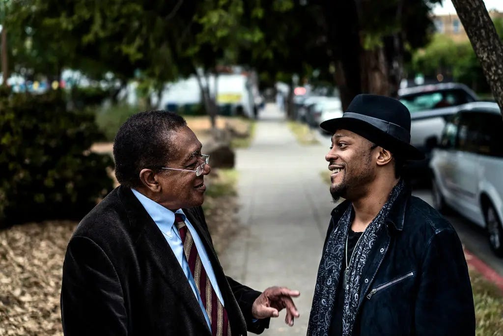 Bobby Seale and D’Angelo standing and conversing on a tree-lined city sidewalk. Bobby Seal wears a suit and tie and D'Angelo wears a dark hat and patterned scarf. Parked cars and greenery are visible in the background. Bobby Seale and D’Angelo standing and conversing on a tree-lined city sidewalk. Bobby Seal wears a suit and tie and D'Angelo wears a dark hat and patterned scarf. Parked cars and greenery are visible in the background.