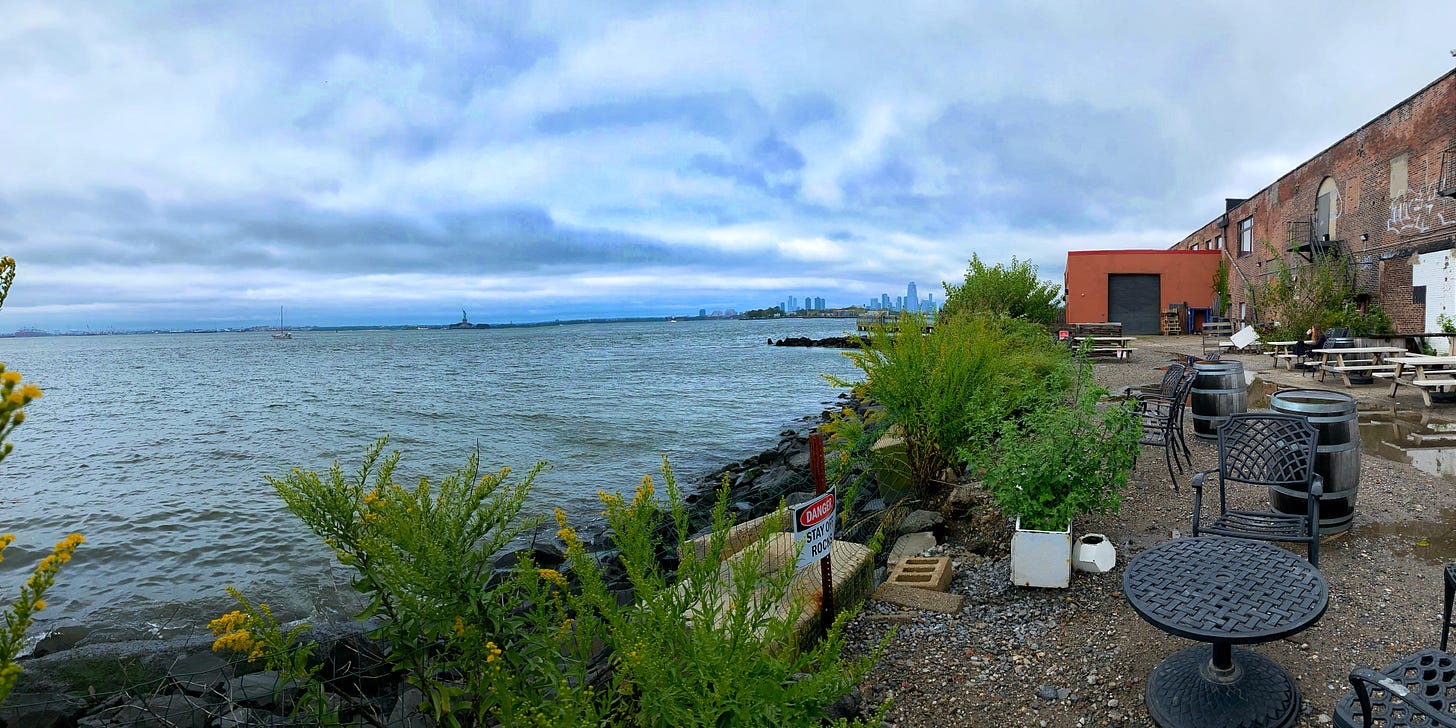 A panoramic photo from deck of Strong Rope Brewery in Red Hook. To the right is an industrial brick building. There are picnic tables in front. Across the water is the city sky line. Further to left, the just clear Statue of Liberty. There is a sailboat on the water, sails furled but mast as high as the buildings with the perspective. To the right are yellow wildflowers alongs the rocks of the shore. A panoramic photo from deck of Strong Rope Brewery in Red Hook. To the right is an industrial brick building. There are picnic tables in front. Across the water is the city sky line. Further to left, the just clear Statue of Liberty. There is a sailboat on the water, sails furled but mast as high as the buildings with the perspective. To the right are yellow wildflowers alongs the rocks of the shore.