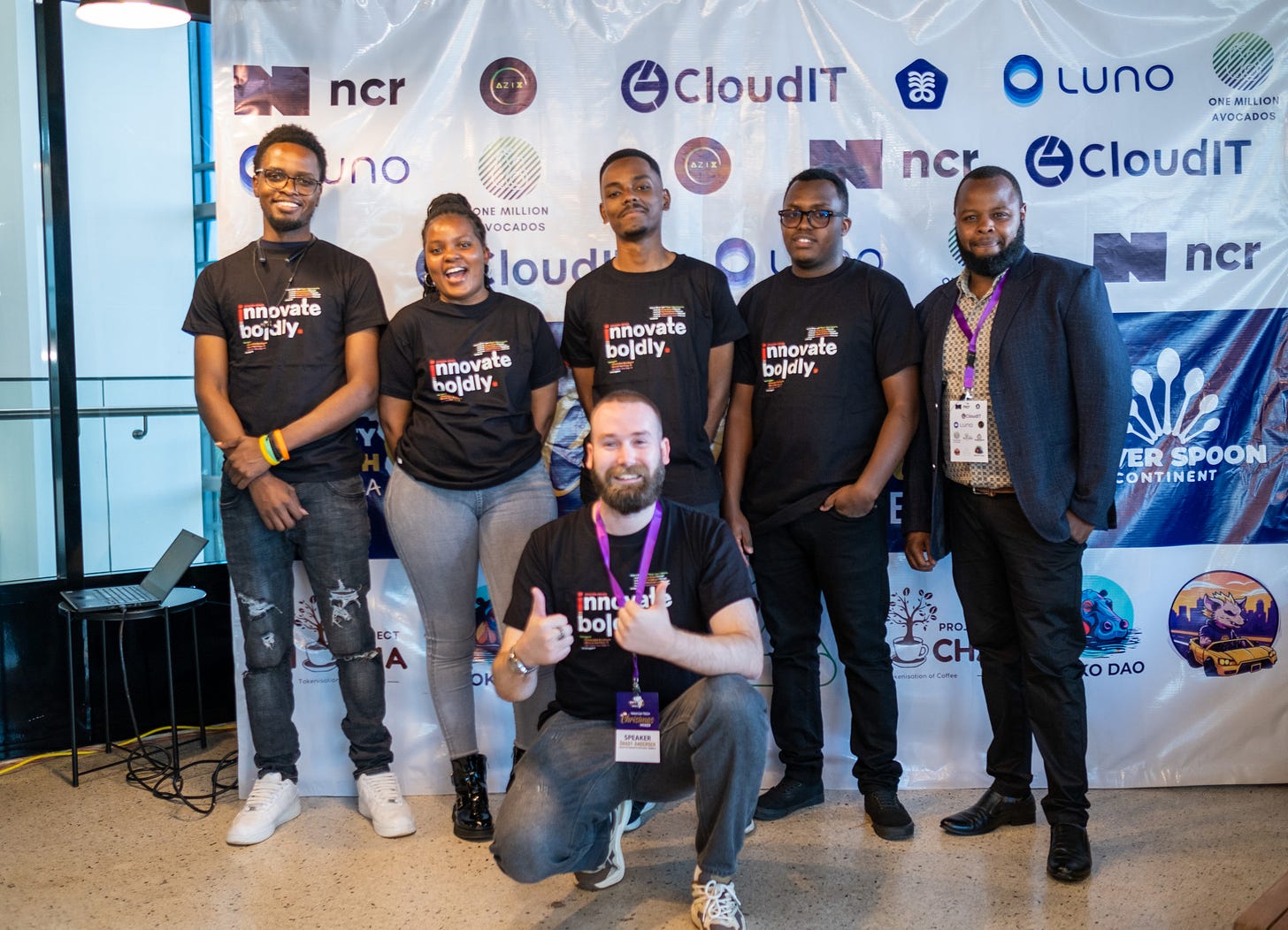 Grady Andersen, founder of nocode rebels, crouches at the front of a group of Kenyan software developers wearing matching 'Innovate Boldly' t-shirts during a tech event in Nairobi. The developers, who are embedded in scaleup teams as part of nocode rebels’s mission, stand smiling in front of a branded backdrop featuring sponsors like CloudIT and Luno.