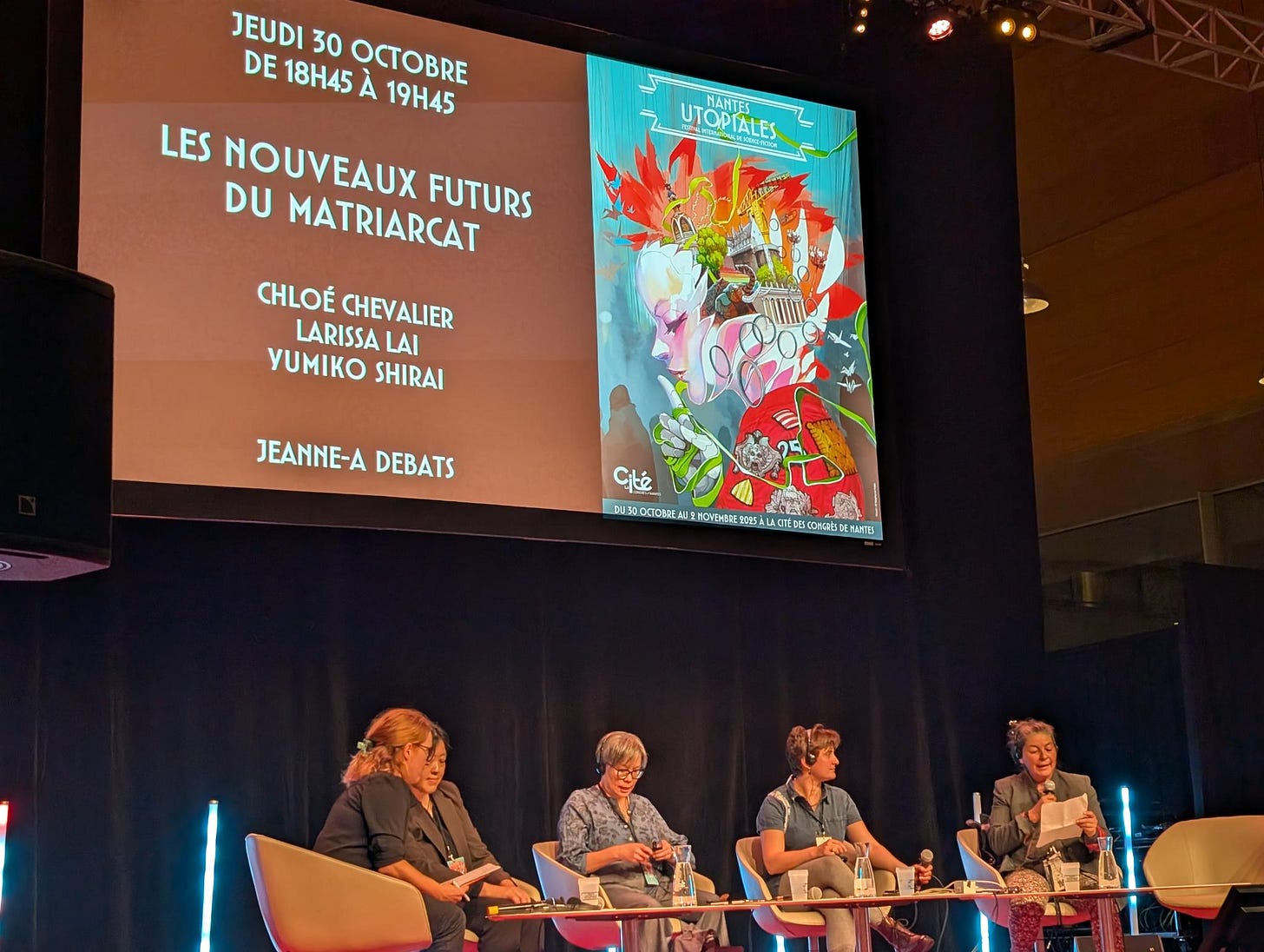 Photographie de la scène de la table ronde, avec les trois participantes, la modératrice et la traductrice d'une des intervenantes. Derrière elles se trouve un écran sur lequel est indiqué le nom de l'évènement et leurs noms. Photographie de la scène de la table ronde, avec les trois participantes, la modératrice et la traductrice d'une des intervenantes. Derrière elles se trouve un écran sur lequel est indiqué le nom de l'évènement et leurs noms.