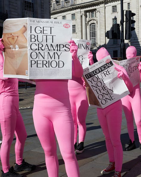 A group of people dressed in bright pink full-body suits holding newspapers titled "The Menstrual Issue," with bold headlines such as "I GET BUTT CRAMPS ON MY PERIOD" and "IT'S OFFICIAL YOU'RE NOT THE ONLY ONE," standing outdoors in an urban setting.  Individuals in bright pink full-body suits sitting and leaning against a building reading newspapers titled "The Menstrual Issue," with bold headlines like "BIG PADS ARE MY HERO."  A group of people in vibrant pink full-body suits holding a newspaper prominently displaying the bold headline "MY PERIOD POOPS ARE CRAZY," outdoors in front of city buildings.