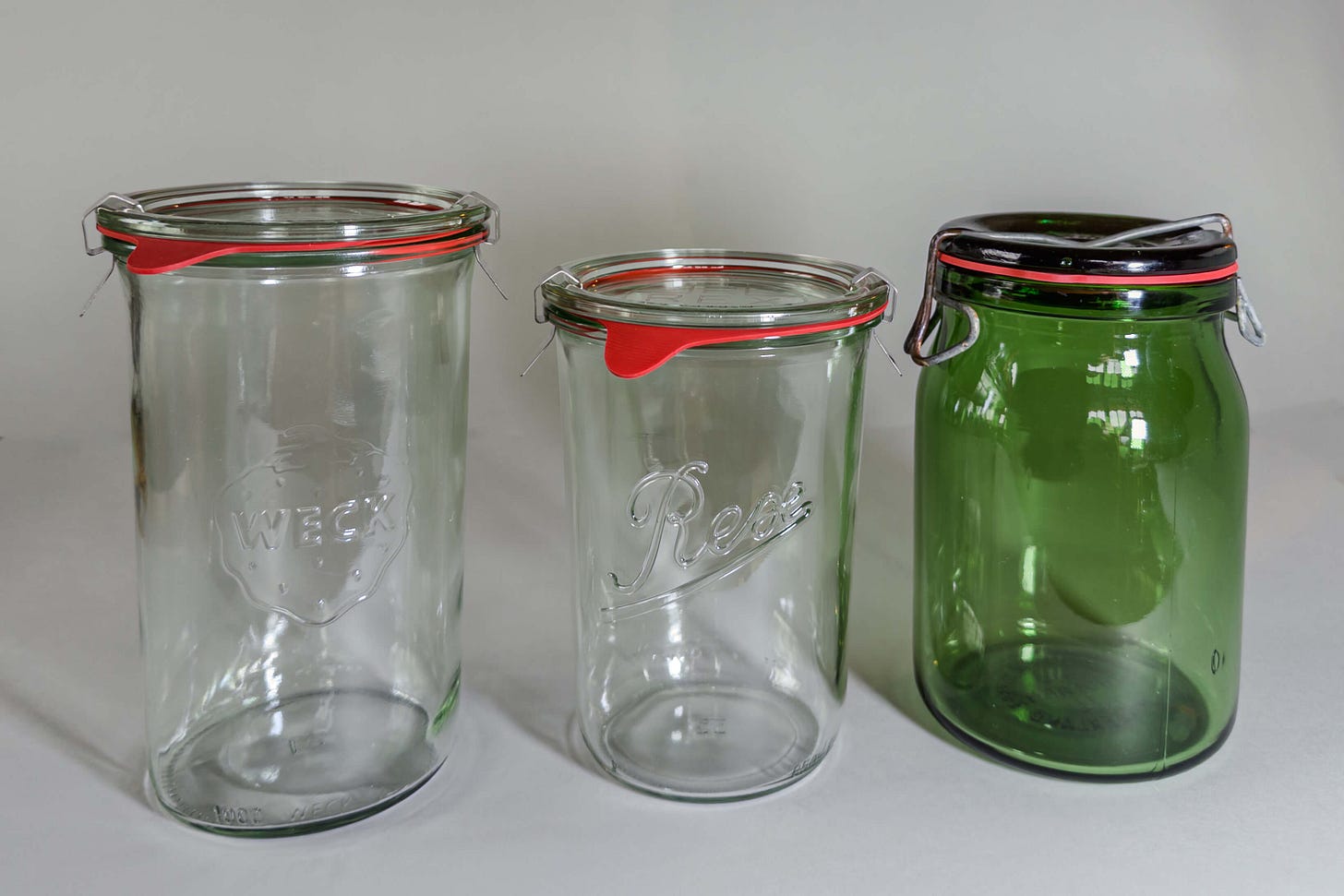 Drei leere Einmachgläser vor hellem Hintergrund: links ein WECK-Glas, in der Mitte ein REX-Glas und rechts ein grünes Bülach-Glas – jeweils mit Glasdeckel, orangefarbenem Gummiring und Metallklemme. (Drei Systeme, eine Aufgabe: das Kraut frisch halten.) Drei leere Einmachgläser vor hellem Hintergrund: links ein WECK-Glas, in der Mitte ein REX-Glas und rechts ein grünes Bülach-Glas – jeweils mit Glasdeckel, orangefarbenem Gummiring und Metallklemme. (Drei Systeme, eine Aufgabe: das Kraut frisch halten.)