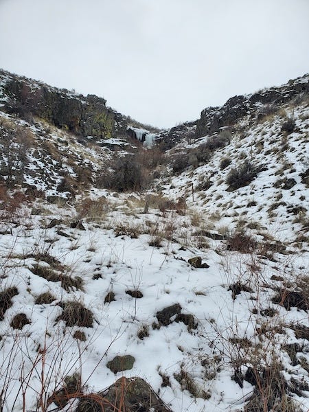looking up a canyon wall in a desert landscape covered by snow, a frozen waterfall at the top of the hill