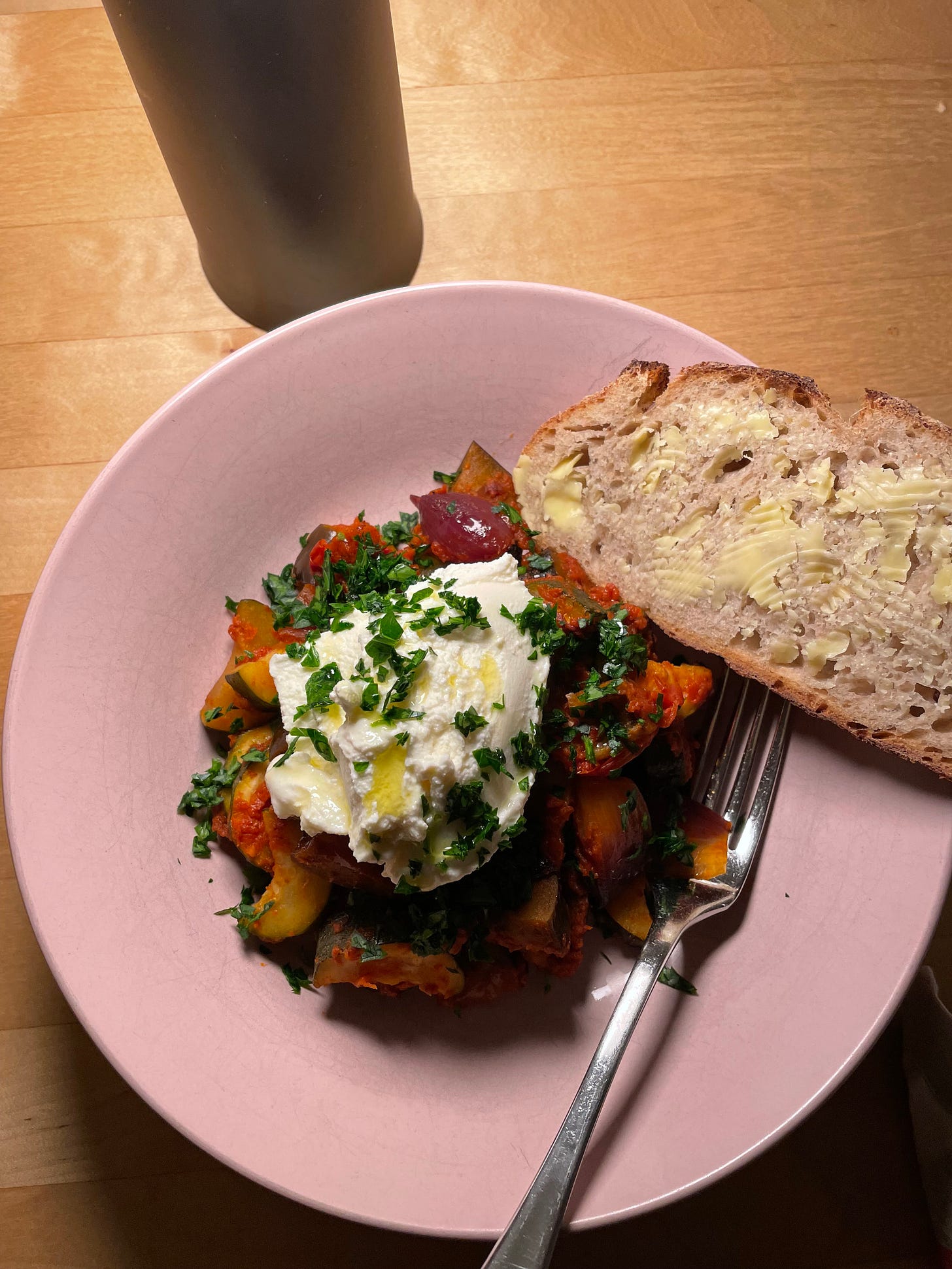 Pink bowl filled with baked ratatouille with red onion, zucchini and eggplant in a rich tomato sauce. It's topped with a scoop of ricotta and chopped parsley and served with a slice of buttered sourdough.