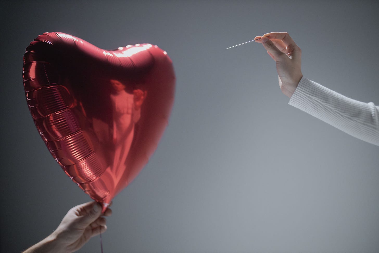 A red heart-shaped foil balloon held up on the left by one hand, while on the right a hand emerging from a white sweater sleeve holds a sewing needle poised to pop it. Grey background.