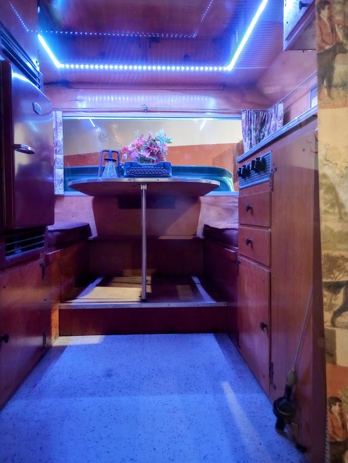 Pickup truck with camper shell, with Rocinante written on side, dog looking out of passenger window, narrow interior with typewriter on table and stove, and man with dog